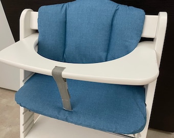May include: A white wooden high chair featuring a blue fabric cushion set for the backrest and seat. The chair includes a smooth white tray and a gray fabric crotch strap attached to the center of the tray, providing a secure seating area.