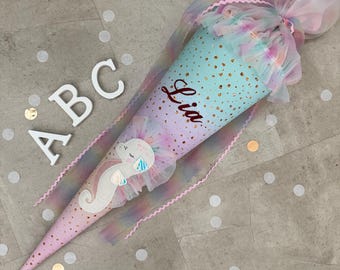May include: A pastel-colored school cone with a seahorse design and the name "Lia" written on it. The cone is decorated with gold dots and tulle, and the letters "ABC" are visible nearby. The cone is designed for school.