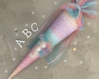 May include: A pastel pink, blue, and gold school cone with a unicorn design and the word "hello" embroidered on it. The cone is wrapped in a sheer pink tulle with white polka dots.