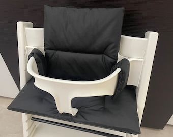 Puede incluir: Silla alta blanca con asiento y respaldo acolchados negros. La silla tiene una bandeja extraíble y un reposapiés.