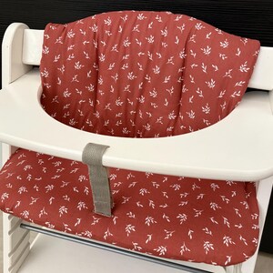 Peut inclure: Chaise haute en bois blanc avec assise et coussin de dossier rouges. Les coussins sont ornés de petites feuilles blanches. Une sangle grise est attachée au siège. La chaise haute est conçue pour un bébé ou un tout-petit.