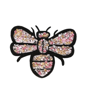Könnte beinhalten: Eine schwarze Filz-Bienenapplikation mit rosa und goldenen Strasssteinen. Die Biene hat einen detaillierten Körper und Flügel.