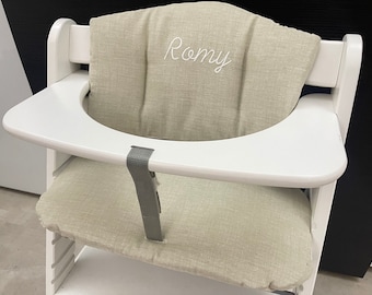 May include: A white wooden high chair featuring a beige fabric cushion set. The back cushion is embroidered with the name Romy in white script. A white tray is attached to the front, and a gray fabric strap is secured to the seat base.
