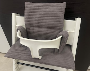 Puede incluir: Una silla alta blanca con un cojín y reposabrazos grises con textura de gofre. La silla tiene una barra de metal negro en la parte delantera.