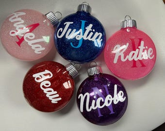 May include: Five round, glittery Christmas ornaments with personalized names: Angela, Justin, Katie, Beau, and Nicole. Each ornament has a silver top.