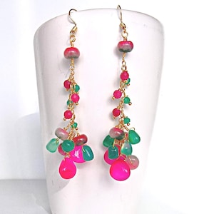 May include: A pair of long earrings with gold-coloured chains and hooks. The earrings are adorned with clusters of pink, green, and red beads and teardrop-shaped stones. They are set against a white backdrop.