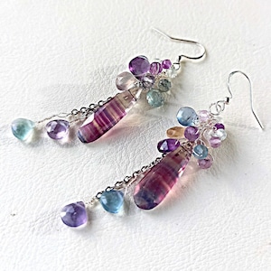Könnte beinhalten: Ein Paar Hängeohrringe mit einem Cluster aus violetten, rosa, blauen und grünen Edelsteinen, die an einer silbernen Kette hängen. Die Ohrringe haben unten einen langen, gestreiften, violetten und rosa Edelstein.