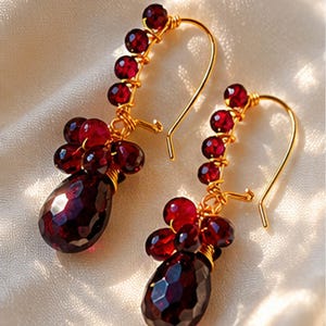 May include: A pair of gold-toned wire-wrapped earrings featuring deep red, faceted garnet beads. Each earring consists of a curved wire stem adorned with a row of small round beads and a cluster of larger, teardrop-shaped stones at the base.