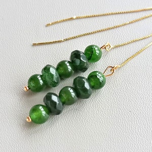 Może przedstawiać: Para zielonych kolczyków z kamieniami szlachetnymi i złotymi łańcuszkami. Kolczyki wykonane są z fasetowanych zielonych kamieni i wiszą na delikatnym złotym łańcuszku.