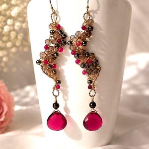 May include: A pair of long dangle earrings featuring a gold-tone wire structure adorned with clusters of small, faceted brown and bright pink beads. Each earring terminates in a single, large, teardrop-shaped, translucent fuchsia crystal drop.