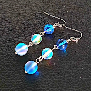 May include: A pair of dangle earrings featuring blue and iridescent glass beads. The earrings have silver hooks and are connected by a silver chain.