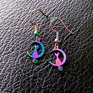 Könnte beinhalten: Ein Paar regenbogenfarbene Ohrringe mit Katze und Mond. Die Ohrringe sind aus Metall und haben eine Regenbogen-Oberfläche.