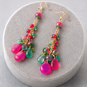 May include: A pair of gold-toned drop earrings featuring clusters of vibrant pink and translucent green teardrop-shaped beads. The beads are suspended from gold-coloured chains attached to standard earring hooks, creating a layered, multi-bead design.