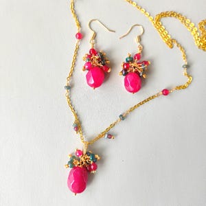 Op de afbeelding: Een gouden sieradenset met een ketting en bijpassende oorbellen. De ketting heeft een hanger met een grote, facetgeslepen roze edelsteen, geaccentueerd door kleinere rode, groene en blauwe kralen. De oorbellen weerspiegelen het hangerontwerp.