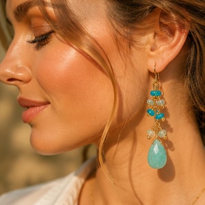 May include: Gold-tone drop earrings featuring turquoise and light blue beads, and a large, teardrop-shaped turquoise stone. The earrings are worn by a person with fair skin and light brown hair.