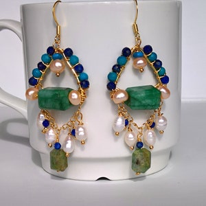 Op de afbeelding: Gouden oorhangers met groene, blauwe en witte kralen. De oorhangers hebben een groene steen, witte parels en blauwe en turquoise kralen.