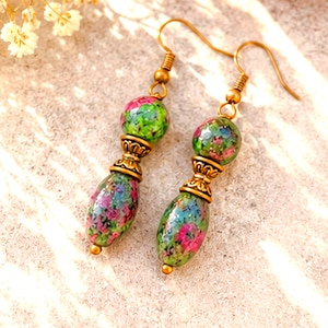 May include: A pair of dangle earrings featuring round and teardrop-shaped beads with a mottled green and magenta pattern. The beads are separated by ornate, antique-style brass spacers and hang from simple brass ear wires with a hook closure.