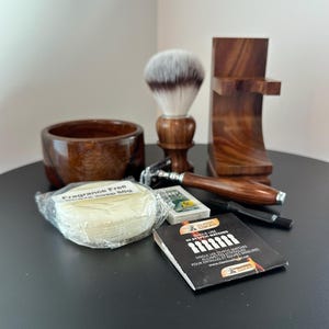 May include: A wooden shaving set with a bowl, brush, razor, and stand. The set includes a bar of fragrance-free shave soap, a pack of styptic matches, and a small box of blades. The items are arranged on a dark surface.