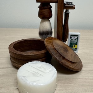 May include: A wooden shaving set with a brush, razor, soap, and a stand. The set is made of dark wood and has a rustic look. The soap is white and is wrapped in plastic. The razor is silver and has a wooden handle. The brush has a white and gray bristle. The stand is made of dark wood and has a simple design.