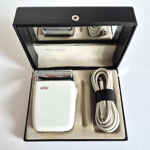 Puede incluir: Una afeitadora eléctrica Braun blanca vintage en un estuche negro. La afeitadora tiene una lámina plateada y el nombre de la marca está impreso en marrón. El estuche incluye un espejo, un cable de carga y una correa negra para sujetar el cable.