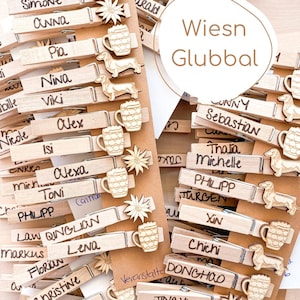 Könnte beinhalten: Holzwäscheklammern mit handgeschriebenen Namen, verziert mit Dackeln, Bierkrügen und Holzblumen. Der Text "Wiesn Glubbal" befindet sich in einem weißen Kreis. Ideal für ein Bastelprojekt oder eine Party.