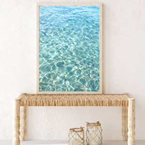 May include: A framed photograph of a turquoise blue ocean with ripples and sunlight reflecting on the surface. The photograph is hanging above a light brown wicker bench with two glass lanterns on the floor in front of it.