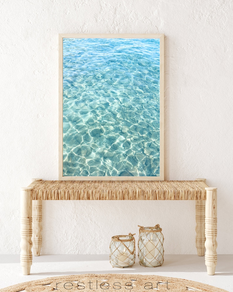 May include: A framed photograph of a turquoise blue ocean with ripples and sunlight reflecting on the surface. The photograph is hanging above a light brown wicker bench with two glass lanterns on the floor in front of it.