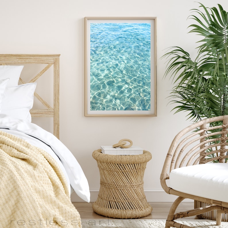 May include: A framed photograph of a turquoise blue ocean with ripples and sunlight reflecting on the surface. The photograph is hanging on a white wall in a bedroom setting.