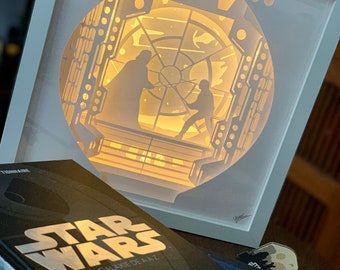 Könnte beinhalten: Gerahmtes, geschichtetes Papierkunstwerk, das eine Szene aus Star Wars mit Darth Vader und Luke Skywalker darstellt. Das Kunstwerk wird von hinten beleuchtet und erzeugt einen warmen Schein. Ein Star Wars-Buch ist im Vordergrund.