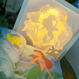 Könnte beinhalten: Ein geschichtetes Papierkunstwerk mit Silhouetten von Arielle und anderen Charakteren aus Arielle, die Meerjungfrau, von innen beleuchtet. Das Kunstwerk ist in einem hellblauen Rahmen gehalten, mit einem geöffneten Kinderbuch im Vordergrund.