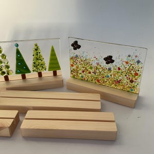 May include: Glass art panels showing trees and a floral scene, set on wooden bases. The tree panels have green and brown details. The floral panel features colourful dots and flower designs. Several additional wooden bases are also visible.