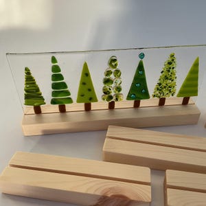 May include: A decorative glass panel featuring six stylized green Christmas trees with brown trunks, set on a light wooden base. The trees vary in design, with some having layered or dotted textures. Additional wooden display stands are visible.