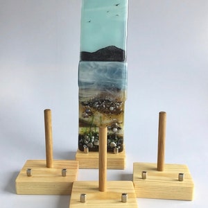 May include: Four wooden display stands with metal accents and a wooden dowel in the centre. Each stand has a square base and is designed to hold a piece of glass art. The glass art is a layered piece with a blue sky, a dark mountain, and a beach scene.