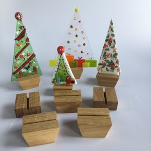 May include: A selection of festive Christmas tree decorations. The designs include a green tree with red details, a clear tree with colourful spots, and a white tree with holly. Each tree is mounted on a wooden base, with additional wooden holders.