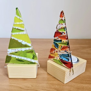 May include: Two decorative glass Christmas tree candle holders. One tree is green and yellow, the other is multi-coloured. Both are set in small, natural wood blocks. The candle holders are on a wooden surface.