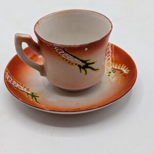 May include: A porcelain teacup and saucer set featuring a cream-colored interior and a vibrant orange exterior rim. The design includes hand-painted dragon motifs in black, yellow, and white, with a curved handle on the side of the cup.