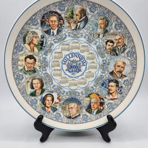 May include: A round ceramic decorative plate featuring a 2000 Millennium Art & Music calendar in the center. The border displays twelve circular portraits of historical figures with blue ornate scrollwork patterns on a white background.