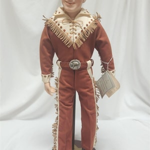 May include: A detailed doll of a man in a Western-style outfit. The doll wears a rust-colored suit with cream-colored accents and fringe. A belt with a silver buckle and a bull design is visible. The doll stands on a wooden base.