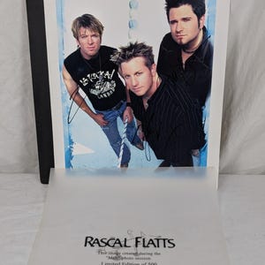 May include: A signed photograph of the musical group Rascal Flatts featuring three members against a blue background. Below the image, a translucent sheet displays the text: RASCAL FLATTS, This image created during the Melt photo session, Limited Edition of 500.