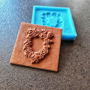 May include: A brown square tile with a floral wreath design is next to a blue silicone mold. The tile has a textured surface and the wreath is raised. The mold is a square shape with the wreath design recessed.