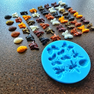 Può includere: Uno stampo in silicone blu a forma di farfalle e api. Ci sono molte farfalle e api di plastica colorate sparse intorno allo stampo.