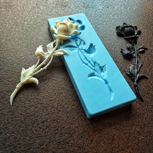 May include: A white rose with a stem and leaves is shown next to a blue silicone mold of a rose with a stem and leaves. The mold is on a brown surface.