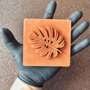 May include: An orange square stamp with a detailed monstera leaf design. The leaf has multiple lobes and small, circular perforations. The stamp is held by a gloved hand, highlighting the intricate carving of the leaf.