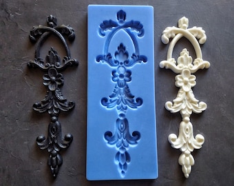 May include: A silicone mold for making decorative cake toppers or other crafts. The mold is blue and features a detailed design of a white, ornate, decorative piece. Two examples of the finished product are shown, one in black and one in white.