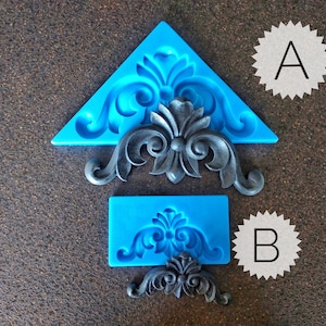 May include: Two blue silicone moulds for making decorative cake toppers. The moulds are shaped like a triangle and a rectangle. The larger mould features a detailed, ornate design. The smaller mould features a simpler design. The moulds are shown with black resin castings of the designs.