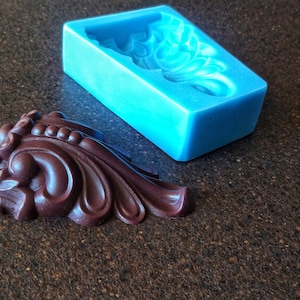 Può includere: Uno stampo in silicone blu e una fusione in cioccolato marrone scuro di un design decorativo e ornato. Lo stampo è rettangolare e la fusione ha la forma di una foglia stilizzata.