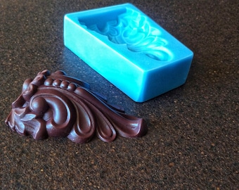 May include: A blue silicone mold and a dark brown chocolate casting of a decorative, ornate design. The mold is rectangular and the casting is shaped like a stylized leaf.