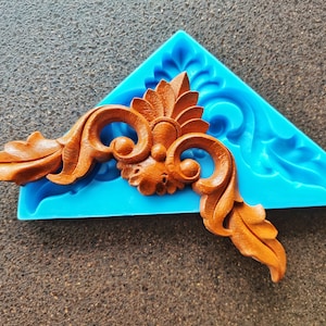 May include: A blue silicone mold with a detailed, intricate, brown wooden design. The design is a corner piece with a floral motif.