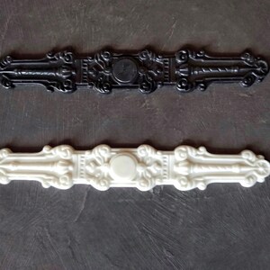 K&ouml;nnte beinhalten: Zwei dekorative Zierleisten, eine schwarze und eine wei&szlig;e, mit verzierten Designs und einem zentralen runden Ausschnitt. Die Zierleisten sind aus Kunststoff und haben eine glatte Oberfl&auml;che.