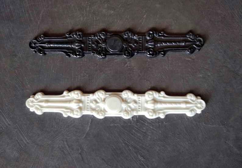 K&ouml;nnte beinhalten: Zwei dekorative Zierleisten, eine schwarze und eine wei&szlig;e, mit verzierten Designs und einem zentralen runden Ausschnitt. Die Zierleisten sind aus Kunststoff und haben eine glatte Oberfl&auml;che.